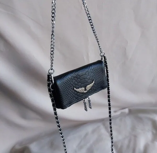Cartera negra con textura de reptil y cadena plateada. Posee un detalle metálico de alas con strass en el frente y dos tiradores colgantes. Ideal para complementar looks de noche o para darle un toque rockero a tu outfit.